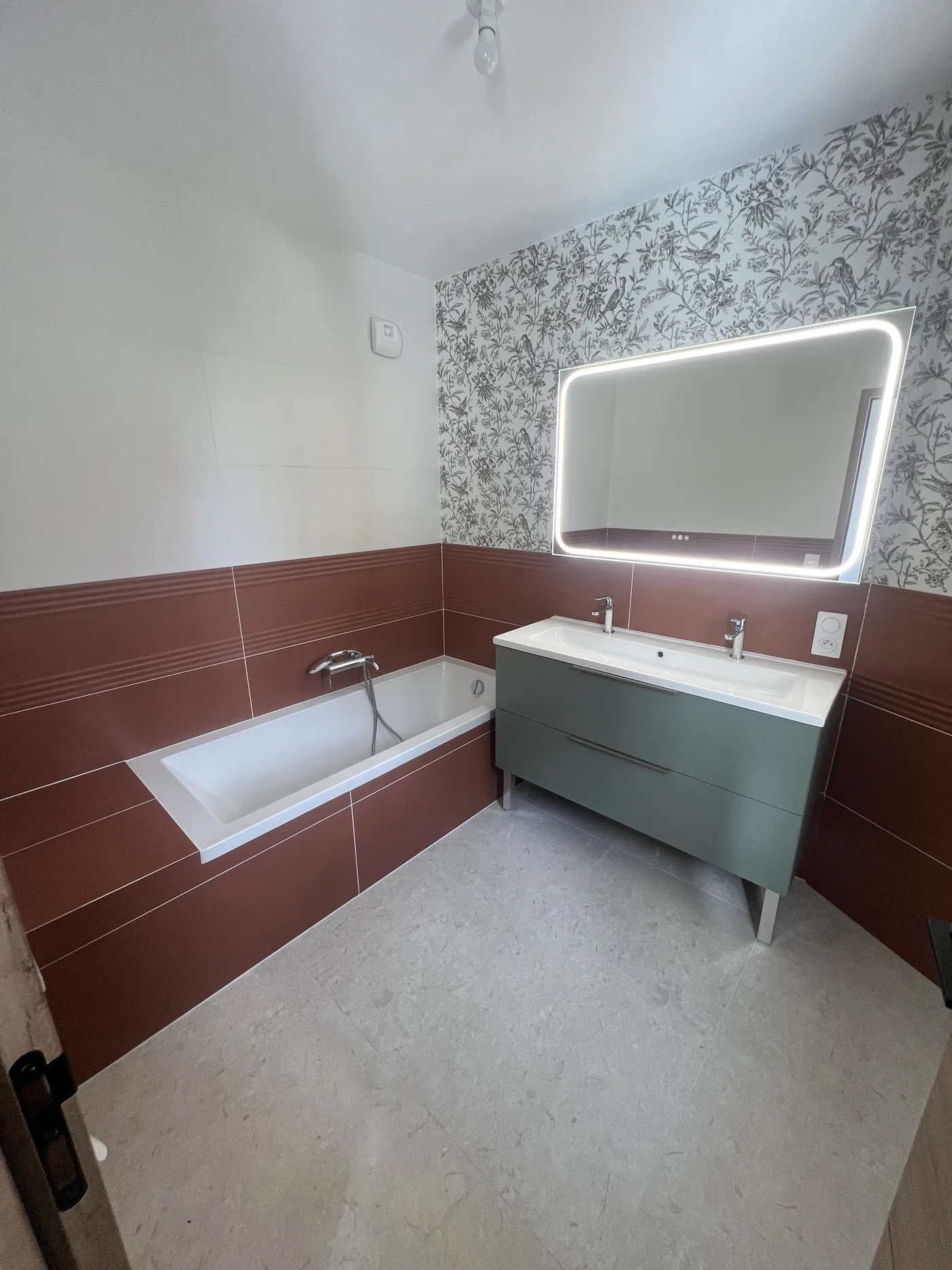 Salle de bain rénovée à Nantes : baignoire encastrée habillée de faïence terracotta, meuble double vasque vert de gris, miroir rétroéclairé et faïence décor à motif fleurs et oiseaux